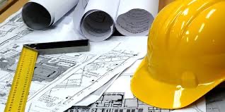 Żółty kask budowlany, kątownik stolarski i zwinięte plany architektoniczne na stole roboczym.