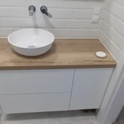 Nowoczesna łazienka z białą umywalką nablatową na drewnopodobnym blacie, białe płytki typu metro na ścianie, kabina prysznicowa z czarną ramą po lewej stronie.