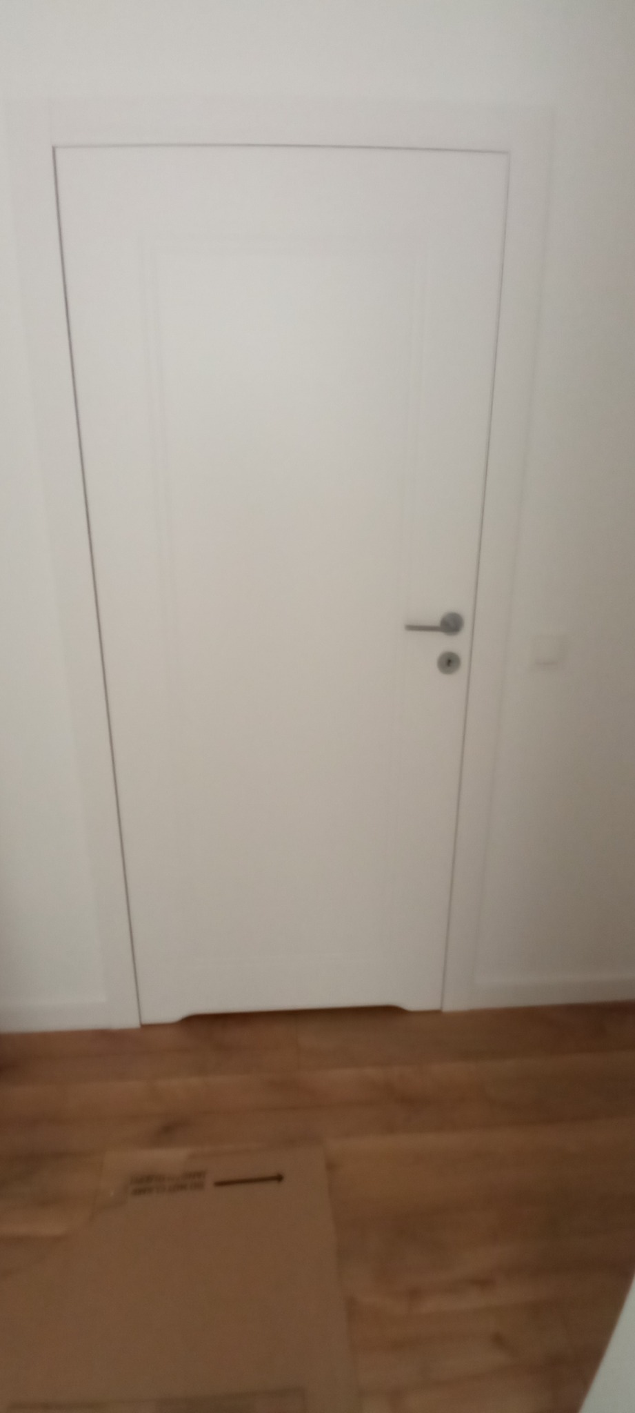 Nowa, biała, minimalistyczna drzwi wewnętrzne z srebrną klamką, widoczne na tle jasnej ściany i drewnianej podłogi z fragmentem kartonu.