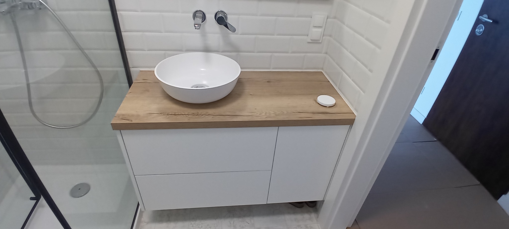 Nowoczesna łazienka z białą umywalką nablatową na drewnopodobnym blacie, białe płytki typu metro na ścianie, kabina prysznicowa z czarną ramą po lewej stronie.