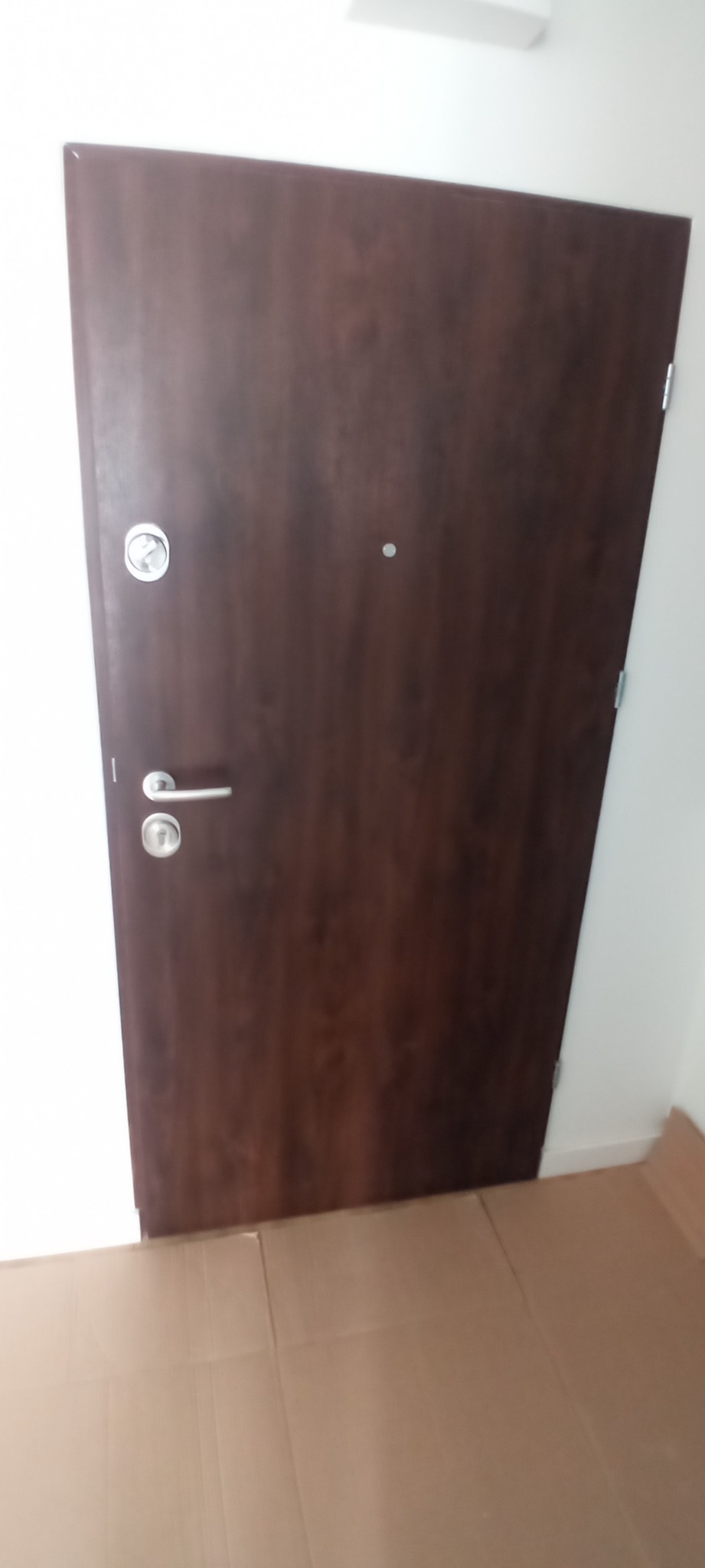 Ciemnobrązowe drzwi wejściowe z srebrną klamką i wizjerem, zabezpieczone kartonem na podłodze, w trakcie lub po montażu.