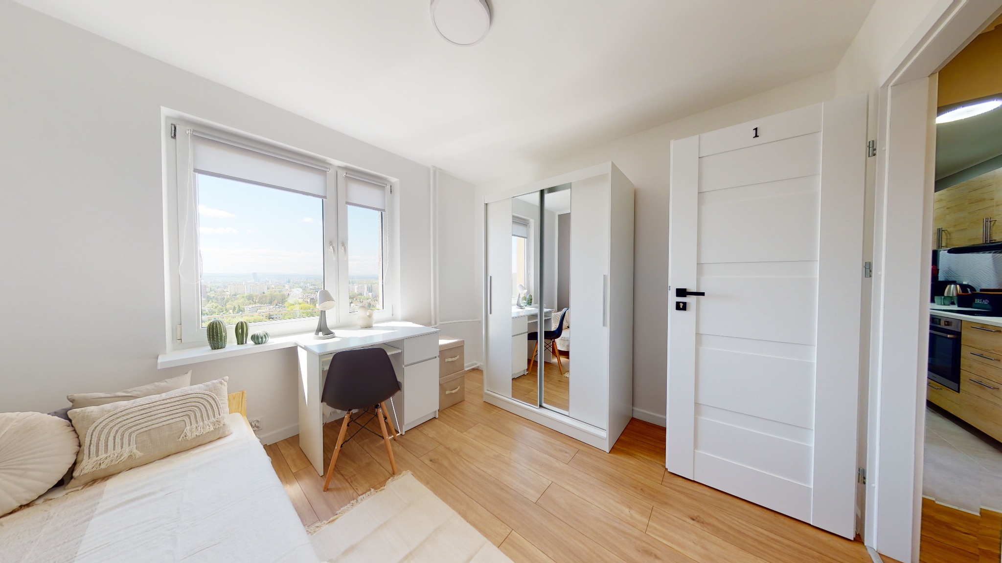 Jasne wnętrze pokoju z biurkiem, łóżkiem i szafą z lustrami, widok na miasto przez okno, drzwi z numerem '1' otwarte na kuchnię.