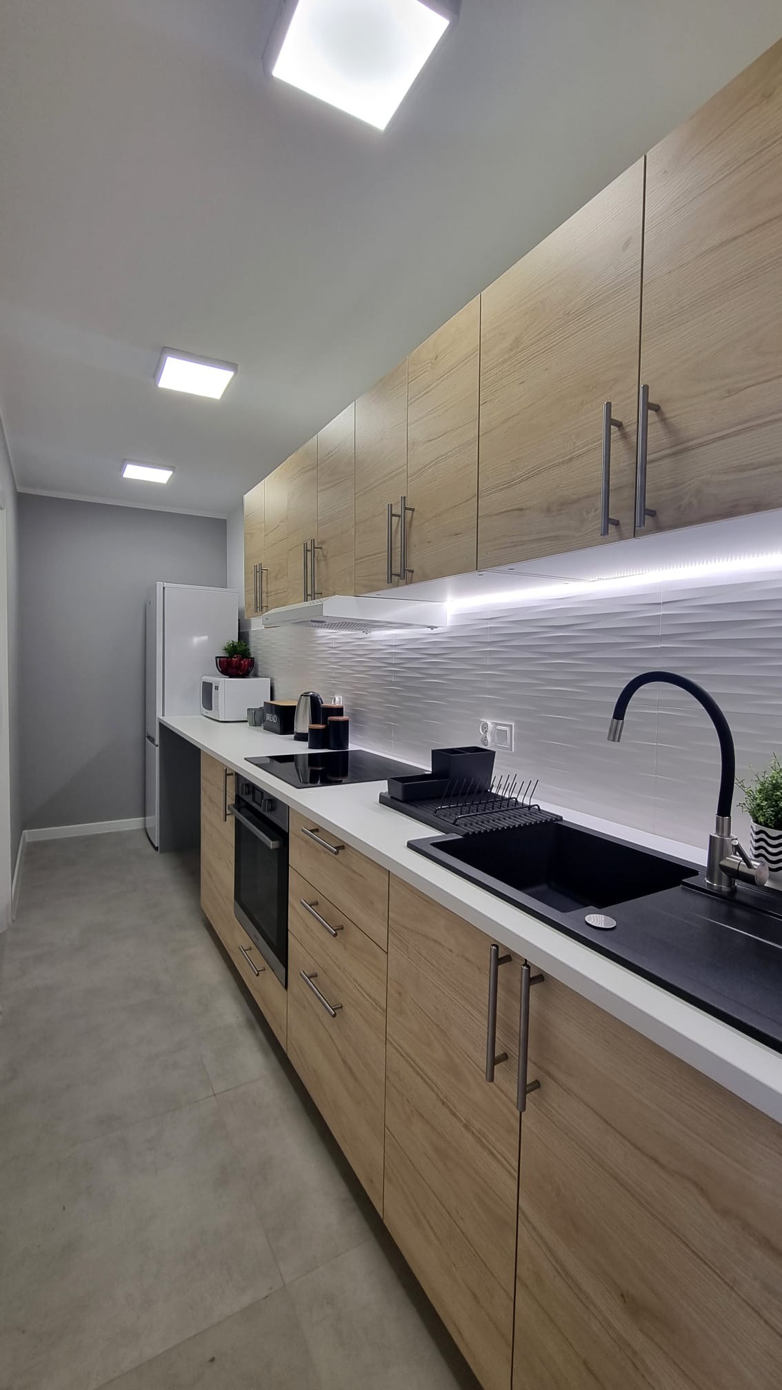 Nowoczesna kuchnia z drewnianymi szafkami, białym blatem i czarnym zlewem, oświetlona ledowymi panelami sufitowymi i listwą pod szafkami, z widocznym drobnym sprzętem AGD.