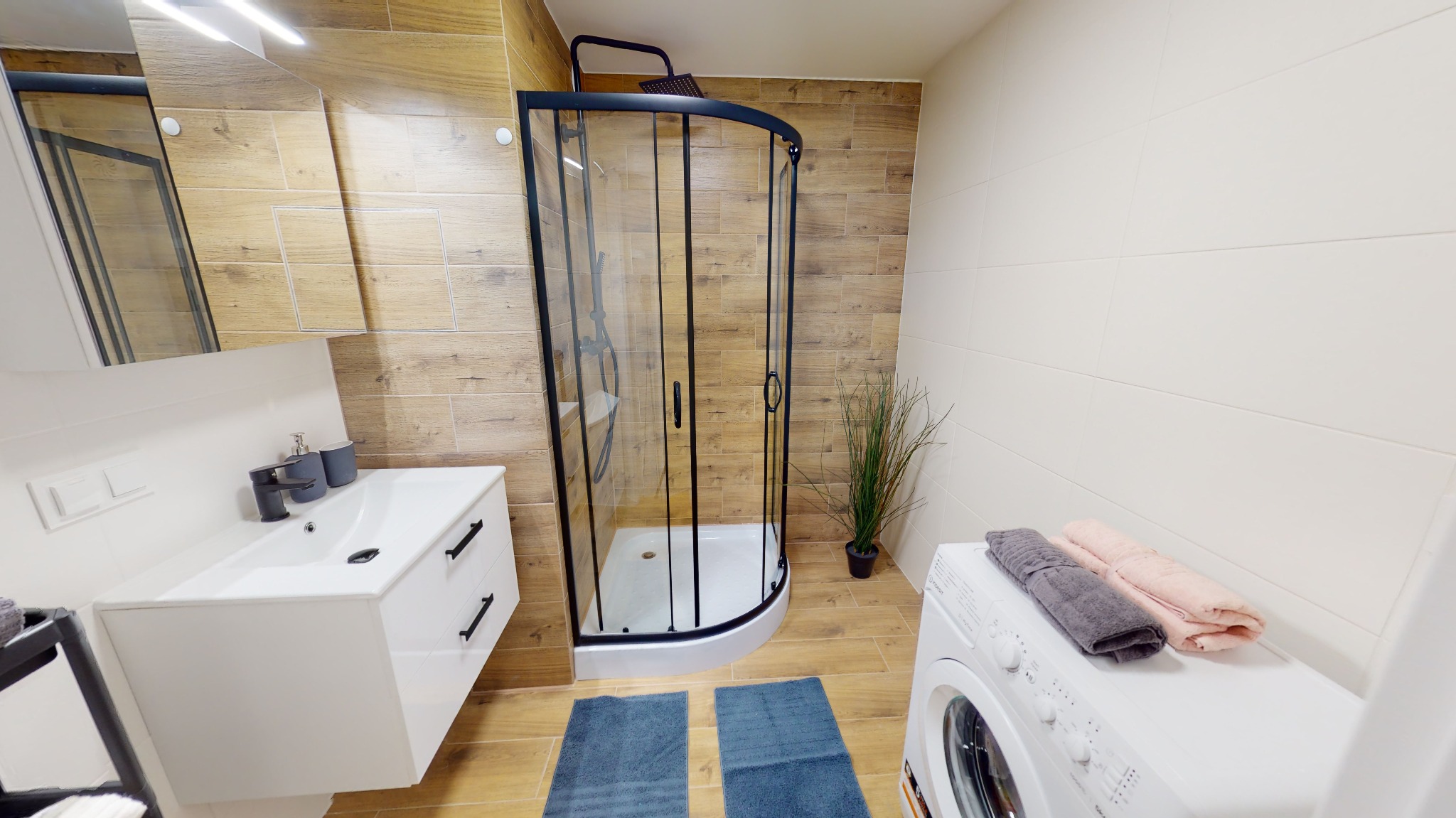 Nowoczesna łazienka z kabiną prysznicową w czarnej ramie, umywalką z szafką, pralką i drewnopodobnymi płytkami na ścianach.