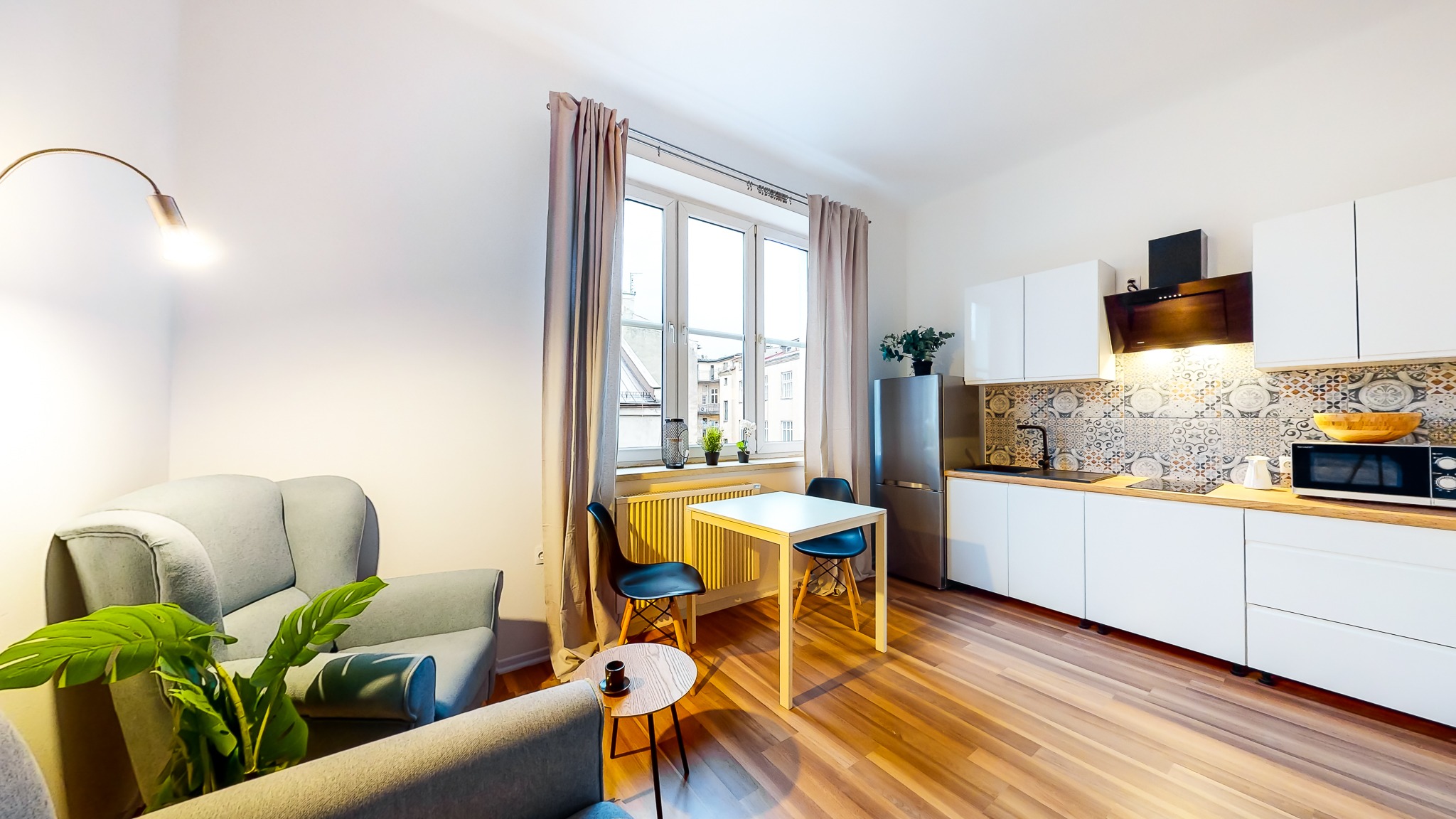 Jasne, nowoczesne studio z aneksem kuchennym, drewnianą podłogą i wygodnym fotelem, oświetlone ciepłym światłem lampy łukowej.