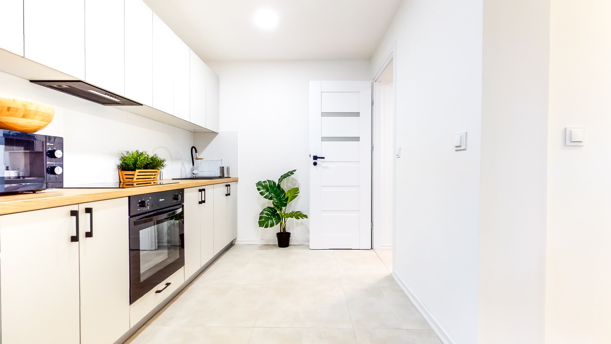 Jasna, minimalistyczna kuchnia z białymi szafkami, drewnianym blatem i czarnymi akcentami, takimi jak uchwyty i piekarnik, z rośliną doniczkową obok białych drzwi z przeszkleniami.
