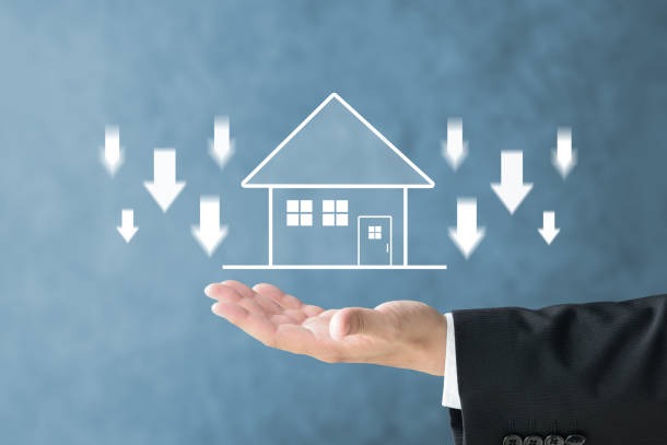 Dłoń w garniturze podtrzymuje schemat domu z białymi strzałkami skierowanymi w dół na rozmytym niebieskim tle, sugerując spadek cen nieruchomości.