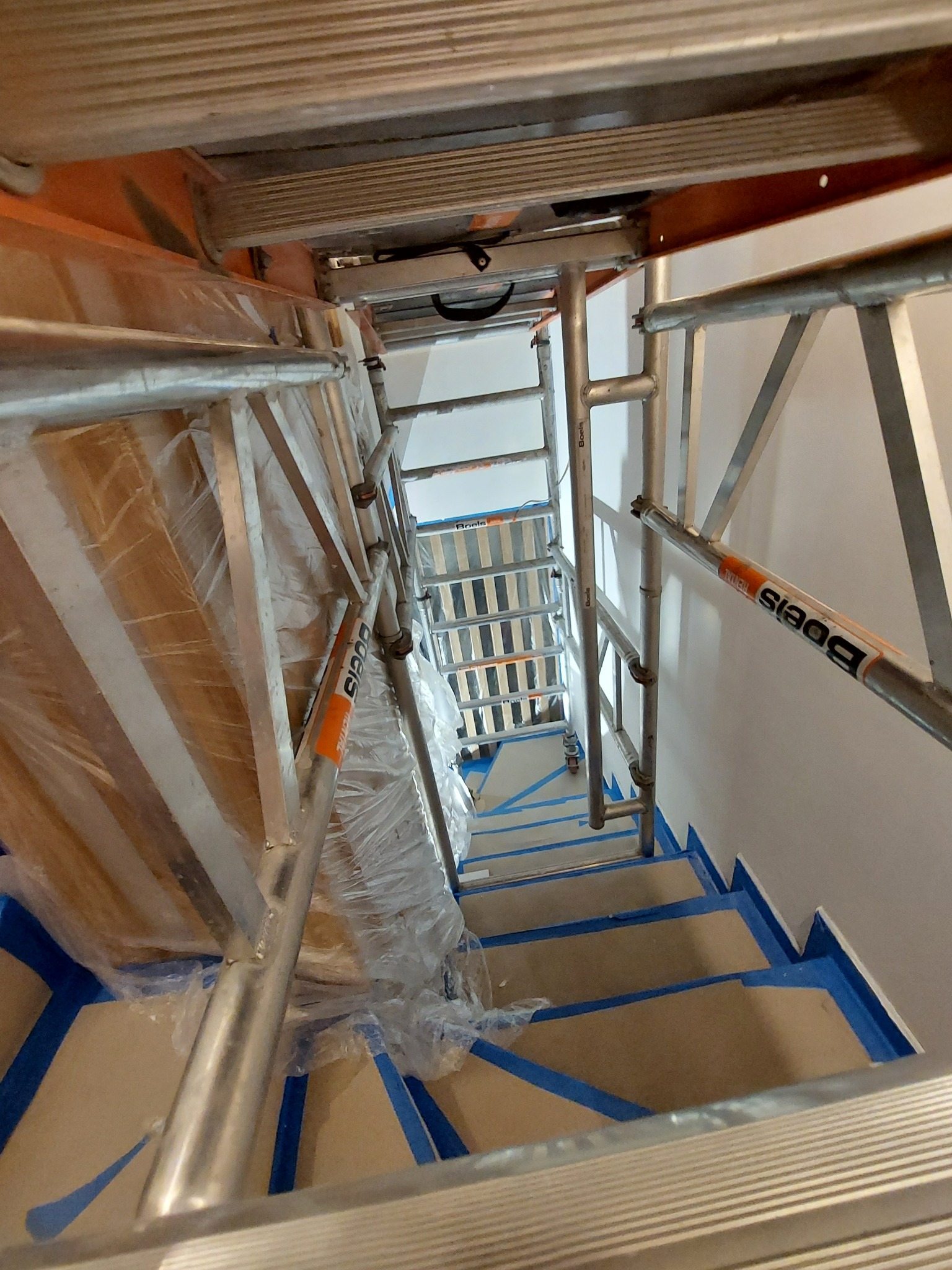 Klatka schodowa zabezpieczona niebieską taśmą malarską, widok z góry na rusztowanie aluminiowe, częściowo owinięte folią, podczas prac wykończeniowych.