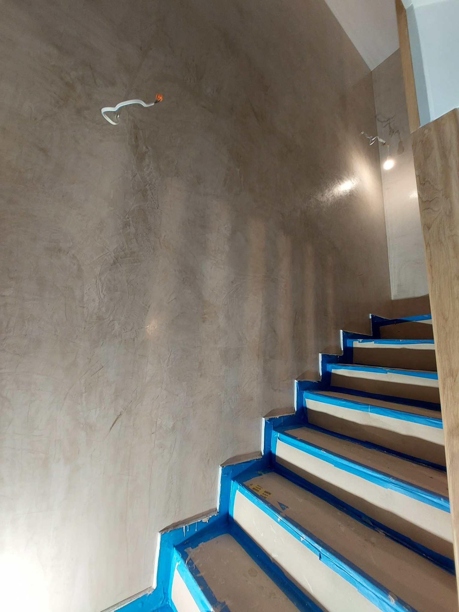 Klatka schodowa w trakcie wykańczania stiukiem, zabezpieczona niebieską taśmą malarską, widoczne przewody elektryczne wystające ze ściany.