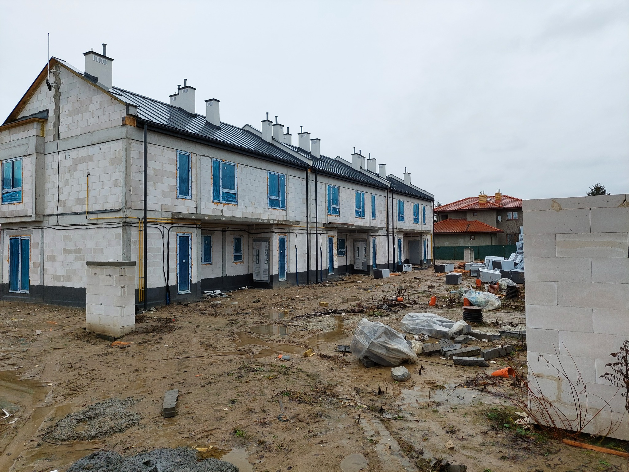 Szeregówka w stanie surowym zamkniętym z pokrytym dachem z blachy, okna i drzwi zabezpieczone folią, teren wokół błotnisty.