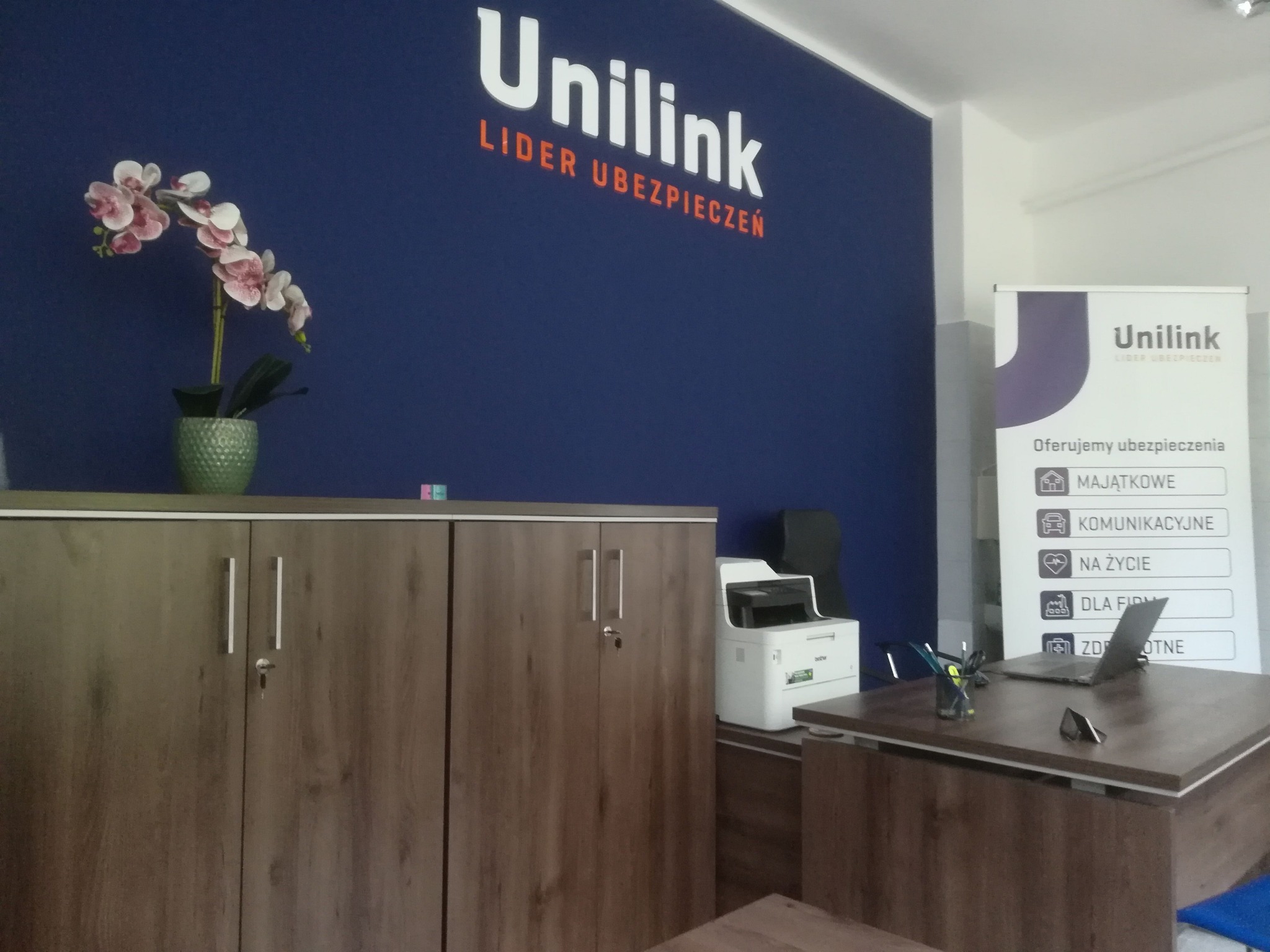 Biuro agencji ubezpieczeniowej Unilink z biurkiem, laptopem, szafką na dokumenty i dekoracyjną orchideą w doniczce.