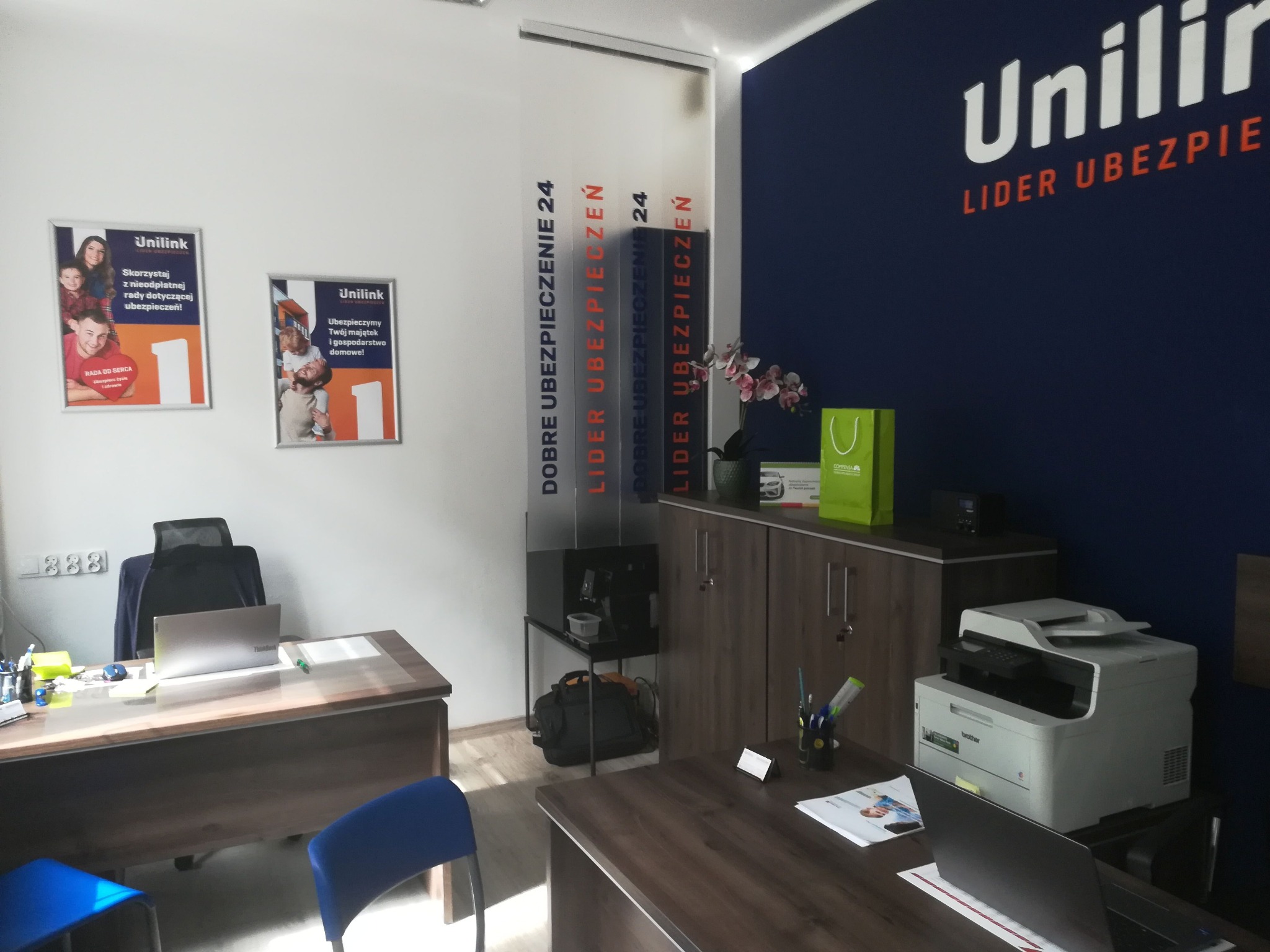 Wnętrze biura firmy ubezpieczeniowej Unilink z dwoma biurkami, laptopami, drukarką Brother, szafką z kwiatami i torbą, plakatami reklamowymi na ścianach i logo firmy na ciemnoniebieskiej ścianie.
