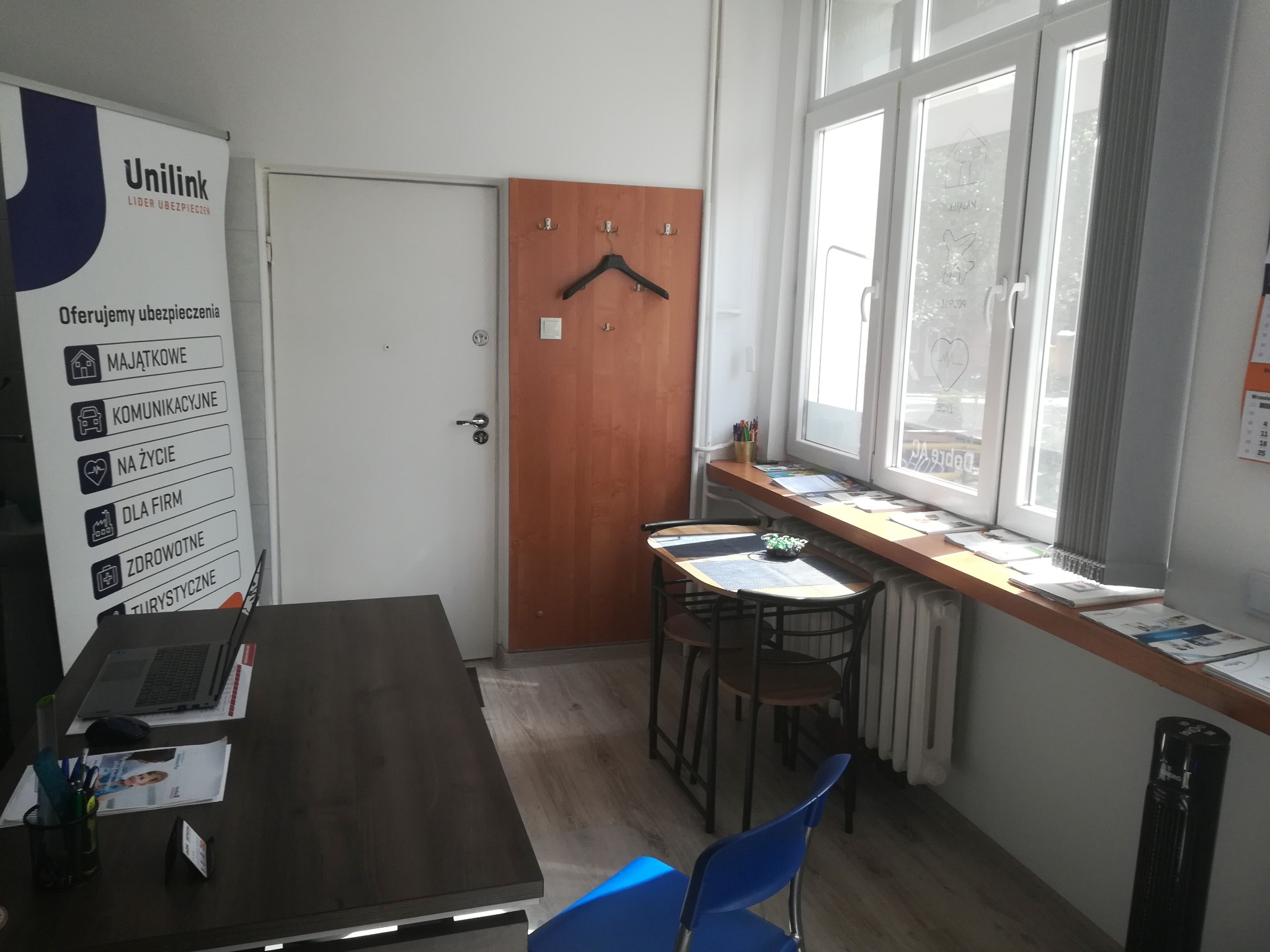 Wnętrze biura ubezpieczeniowego z biurkiem, laptopem, krzesłami i plakatem reklamowym Unilink z ofertą ubezpieczeń majątkowych, komunikacyjnych, na życie, dla firm, zdrowotnych i turystycznych.