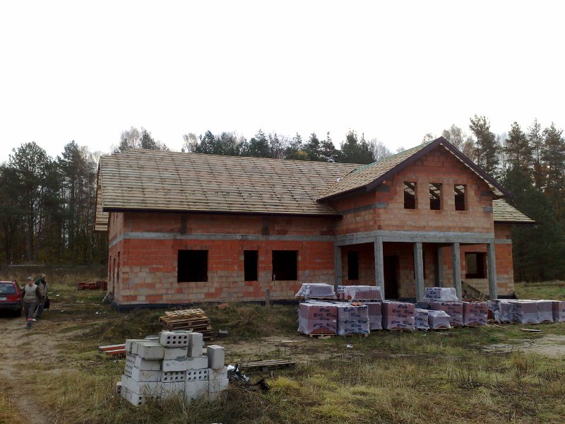 Dom w stanie surowym otwartym w Działdowie z częściowo wykonanym pokryciem dachowym z dachówki ceramicznej, widoczne materiały budowlane na posesji i las w tle.