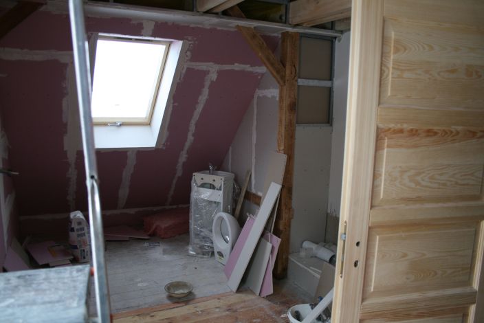 Poddasze w trakcie adaptacji: widoczne okno dachowe, ocieplenie, płyty gipsowo-kartonowe i elementy konstrukcji drewnianej. W tle sanitariaty przygotowane do montażu.