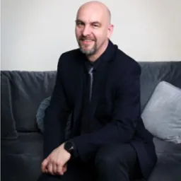 Portret uśmiechniętego mężczyzny w garniturze, siedzącego na szarej sofie z poduszkami, z zegarkiem na nadgarstku.