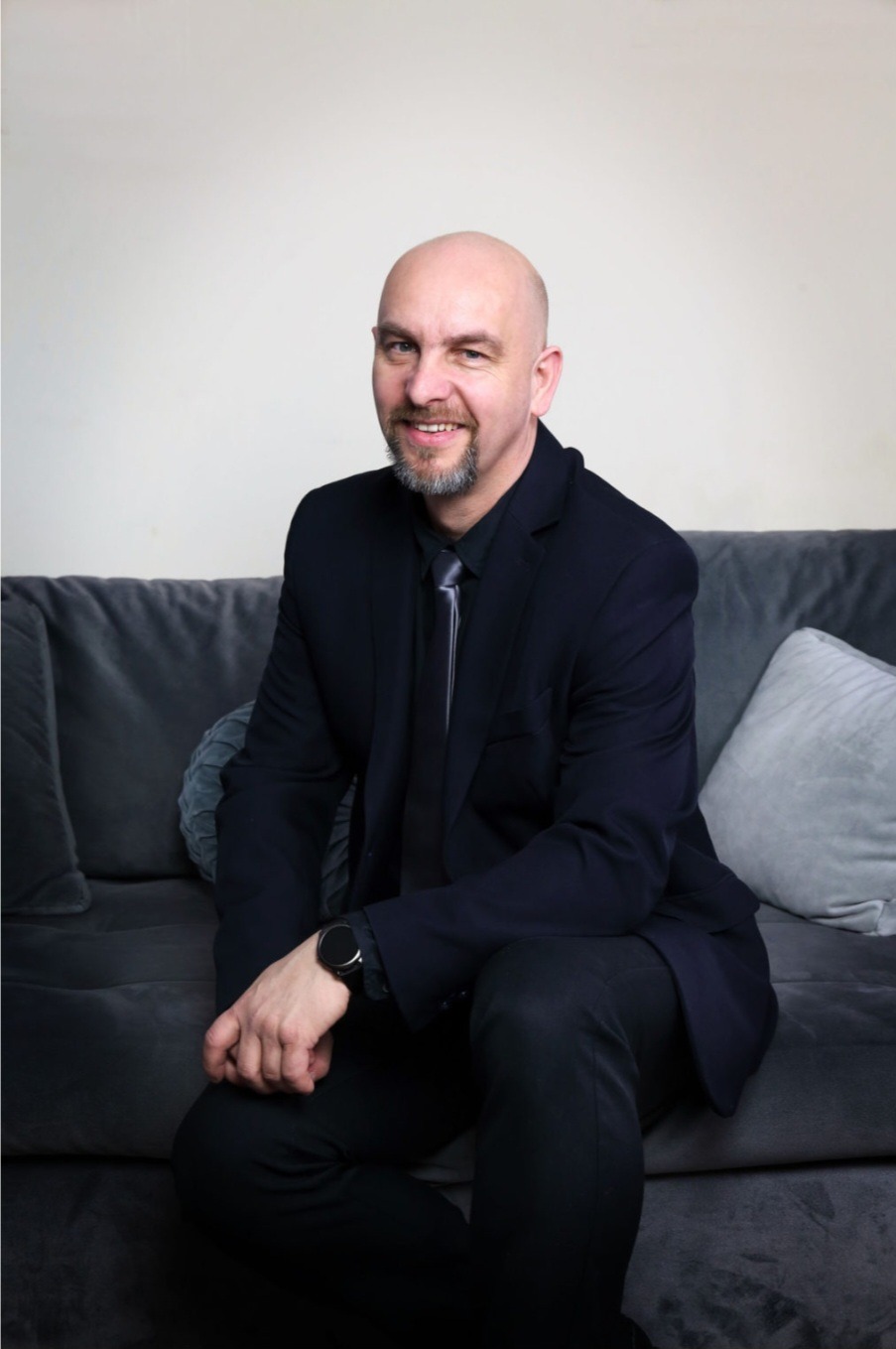 Portret uśmiechniętego mężczyzny w garniturze, siedzącego na szarej sofie z poduszkami, z zegarkiem na nadgarstku.