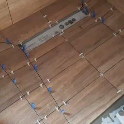 Żarbud - Układanie drewnopodobnych płytek podłogowych w łazience, widoczne krzyżyki dystansowe i niebieskie kliny poziomujące, odpływ liniowy w posadzce.