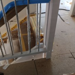 Żarbud - Fragment balustrady schodowej z drewnianym pochwytem zabezpieczonym niebieską pianką, metalowymi prętami w folii ochronnej i pomalowanym na biało słupkiem, widoczne elementy podłogi w trakcie...