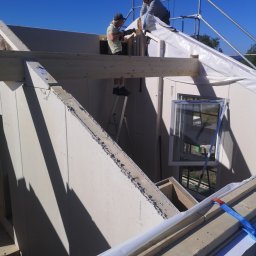 Żarbud - Dwóch mężczyzn pracuje na wysokościach przy montażu konstrukcji dachu domu w słoneczny dzień, jeden na drabinie, drugi na rusztowaniu, widoczne okno w ścianie.