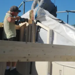 Dwóch mężczyzn pracuje na rusztowaniu przy budowie dachu, jeden wierci w drewnianej konstrukcji, drugi układa izolację z folii, widok na jasne niebo.