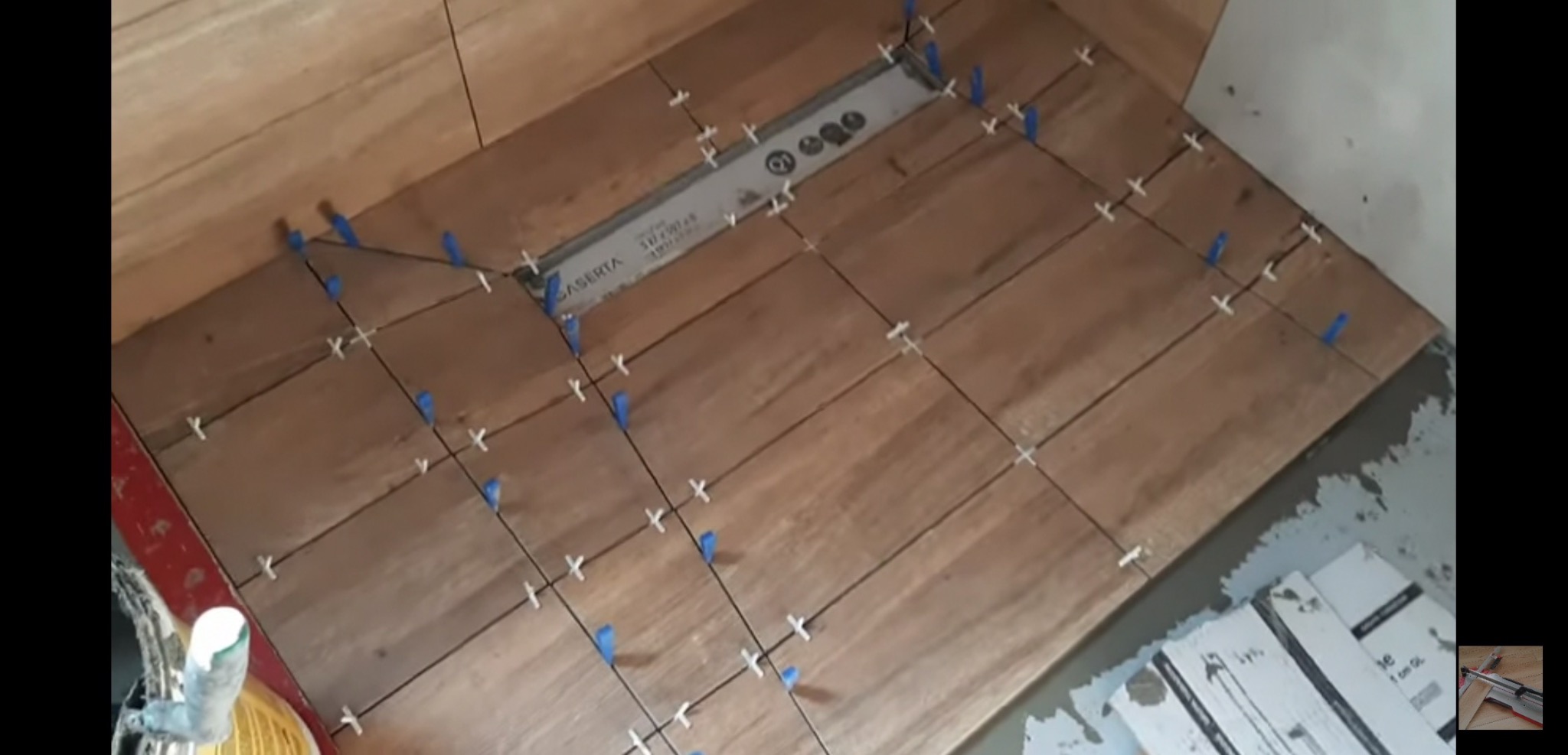 Układanie drewnopodobnych płytek podłogowych w łazience, widoczne krzyżyki dystansowe i niebieskie kliny poziomujące, odpływ liniowy w posadzce.