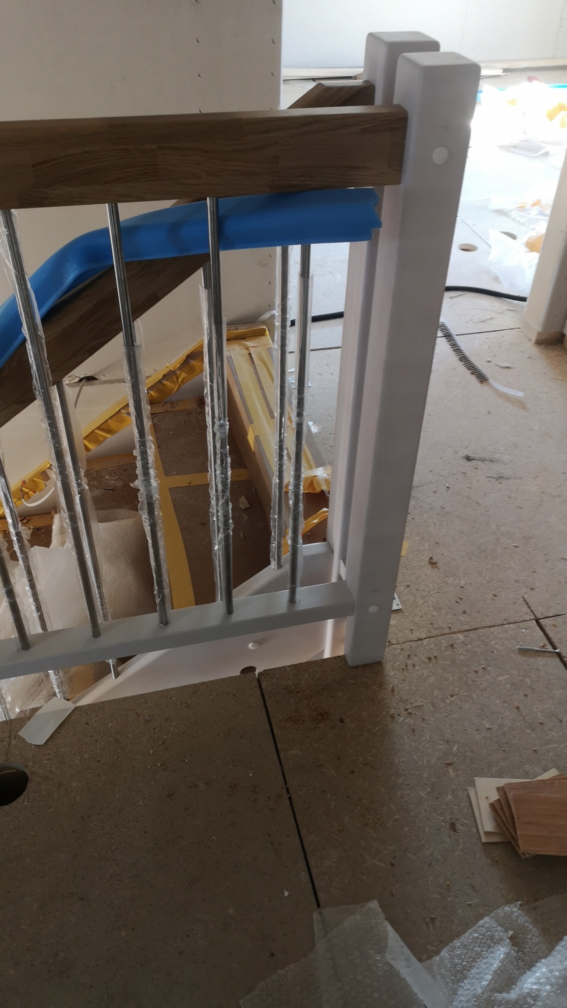 Fragment balustrady schodowej z drewnianym pochwytem zabezpieczonym niebieską pianką, metalowymi prętami w folii ochronnej i pomalowanym na biało słupkiem, widoczne elementy podłogi w trakcie...
