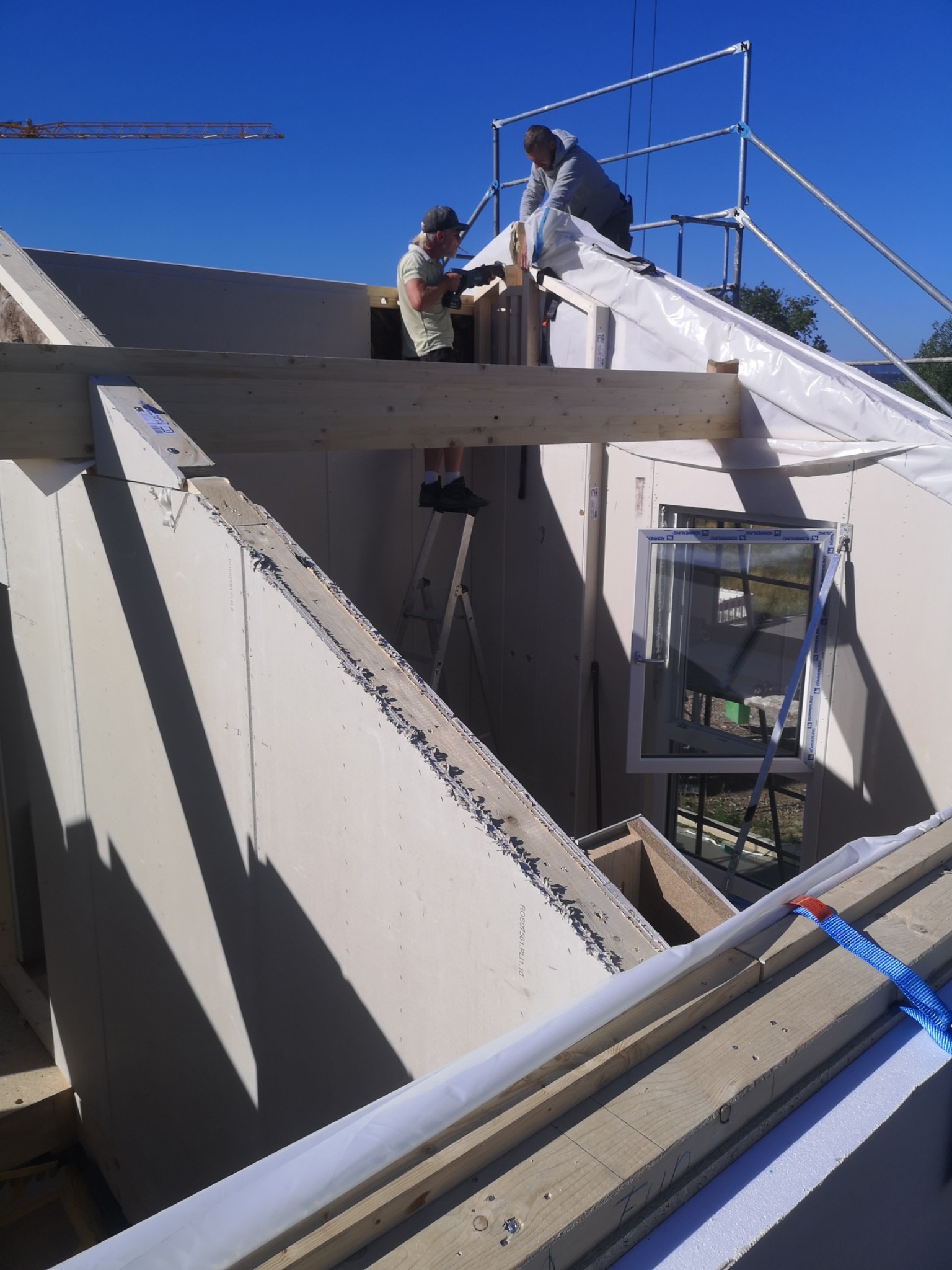 Dwóch mężczyzn pracuje na wysokościach przy montażu konstrukcji dachu domu w słoneczny dzień, jeden na drabinie, drugi na rusztowaniu, widoczne okno w ścianie.