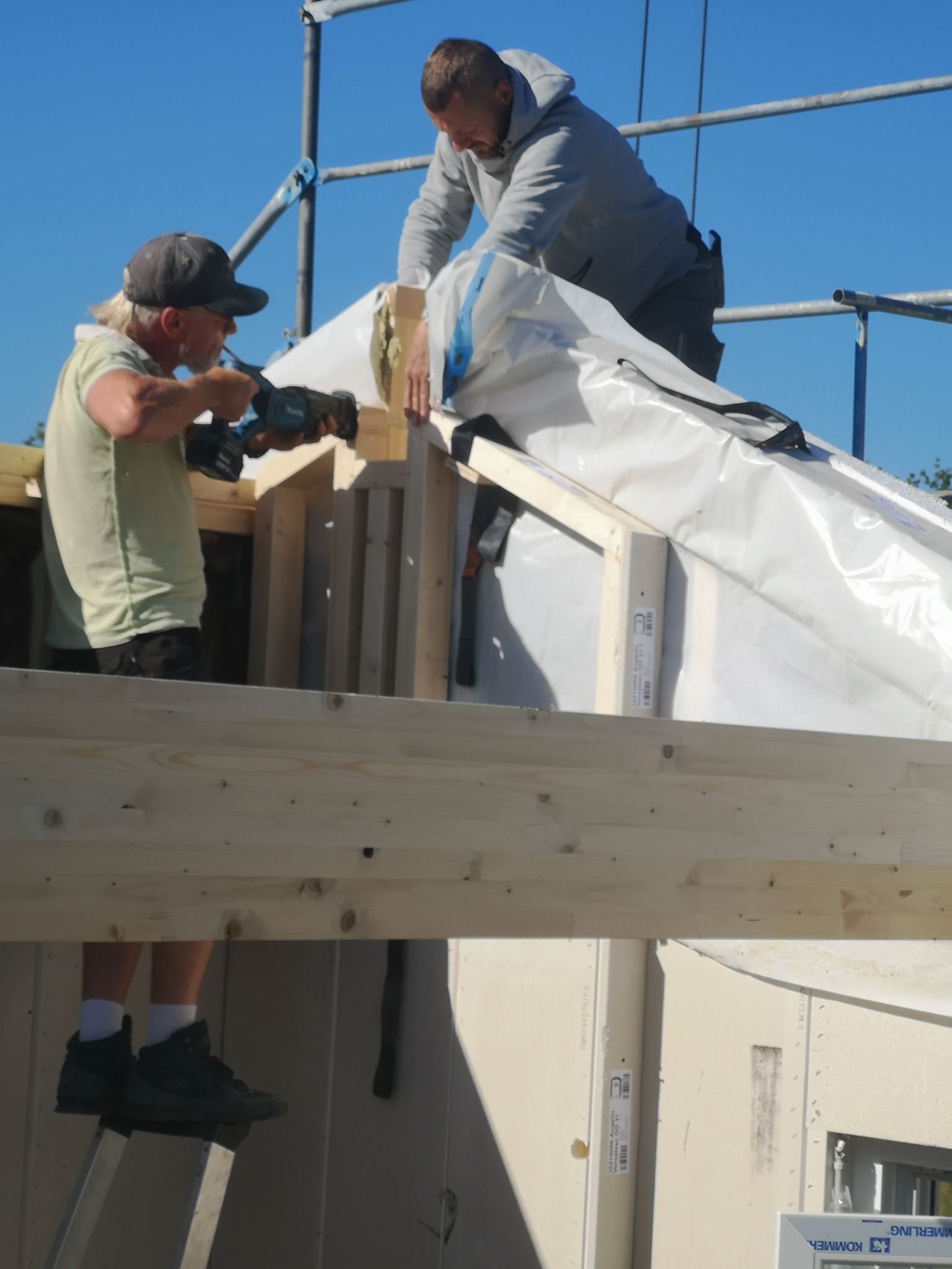 Dwóch mężczyzn pracuje na rusztowaniu przy budowie dachu, jeden wierci w drewnianej konstrukcji, drugi układa izolację z folii, widok na jasne niebo.