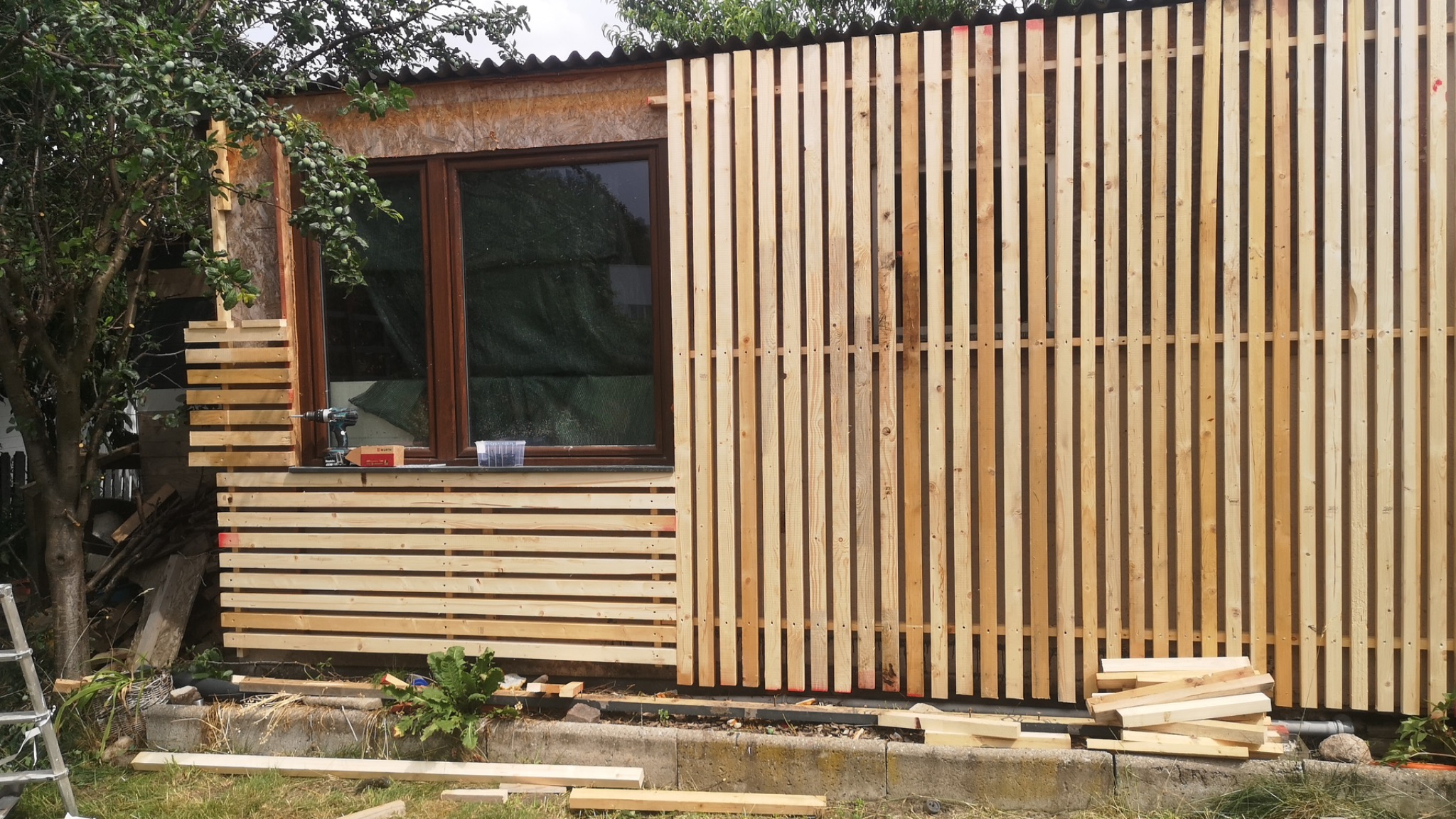 Montaż drewnianej elewacji z pionowych listew na budynku, widoczne okno, wiertarka i rozsypane deski na trawie.