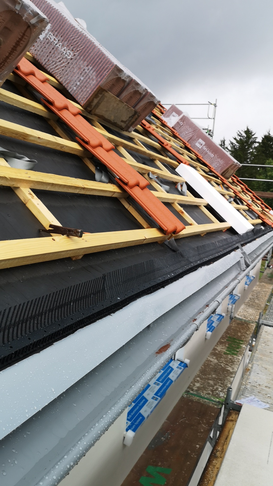 Montaż dachówki ceramicznej na drewnianej konstrukcji dachu, widoczne krokwie, membrana dachowa, paczki dachówek Braas w folii, orynnowanie z elementami montażowymi.