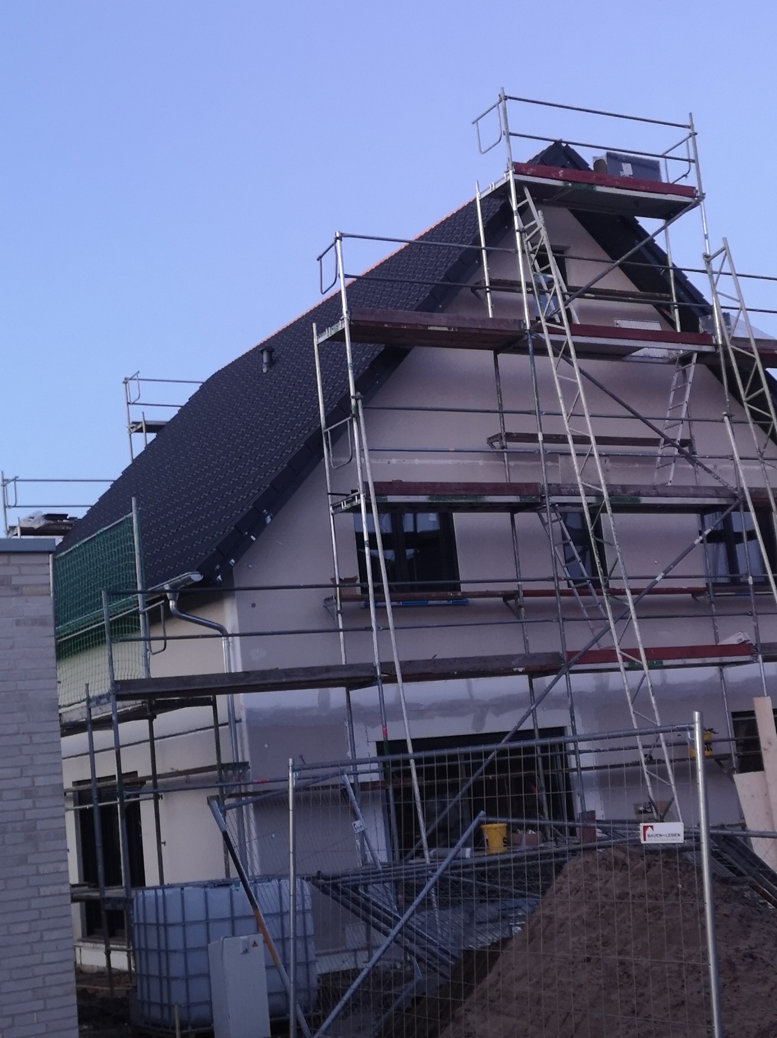 Dom jednorodzinny w trakcie wykończenia elewacji, otoczony rusztowaniami i zabezpieczony siatką, z widocznymi oknami i nowym dachem z dachówki.