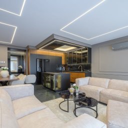 MW Wnętrza - Nowoczesny salon z aneksem kuchennym, z geometrycznym oświetleniem LED na suficie, szarymi ścianami z listwami, beżowymi sofami i czarnymi stolikami kawowymi. Ciemna zabudowa kuchenna z drewnianymi...