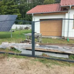 Szara brama panelowa z zaokrąglonym górnym brzegiem, widoczna na tle domu z panelami słonecznymi, w trakcie montażu, z odkrytymi fundamentami.