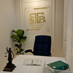 Wnętrze gabinetu prawniczego: biurko z otwartą książką, statuetka Temidy, zielona roślina, logo kancelarii na ścianie, ciemnoniebieskie krzesło.