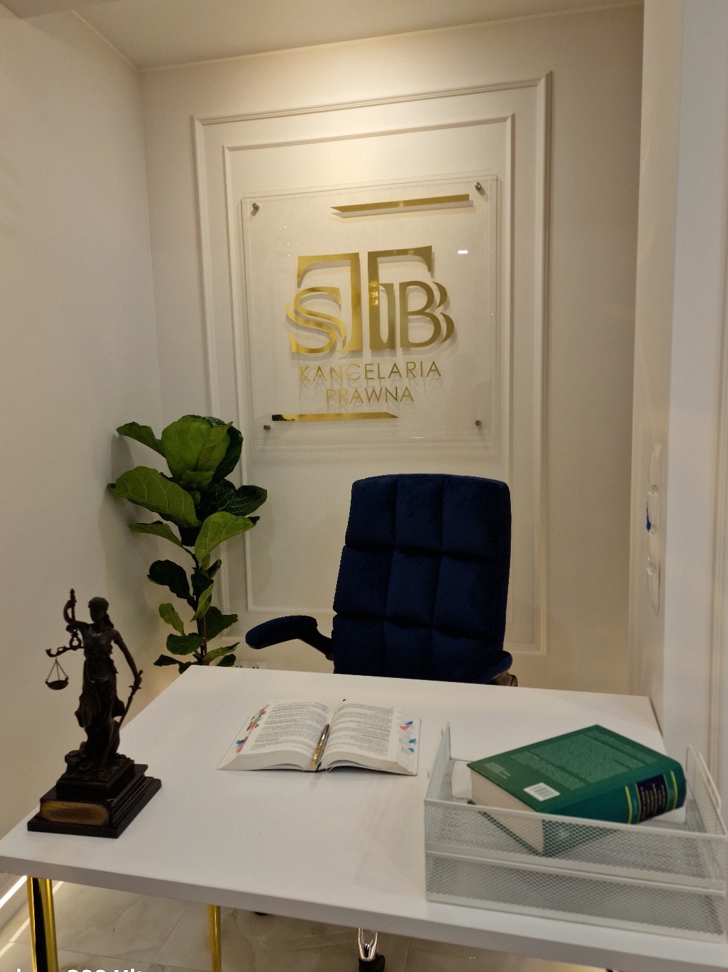 Wnętrze gabinetu prawniczego: biurko z otwartą książką, statuetka Temidy, zielona roślina, logo kancelarii na ścianie, ciemnoniebieskie krzesło.