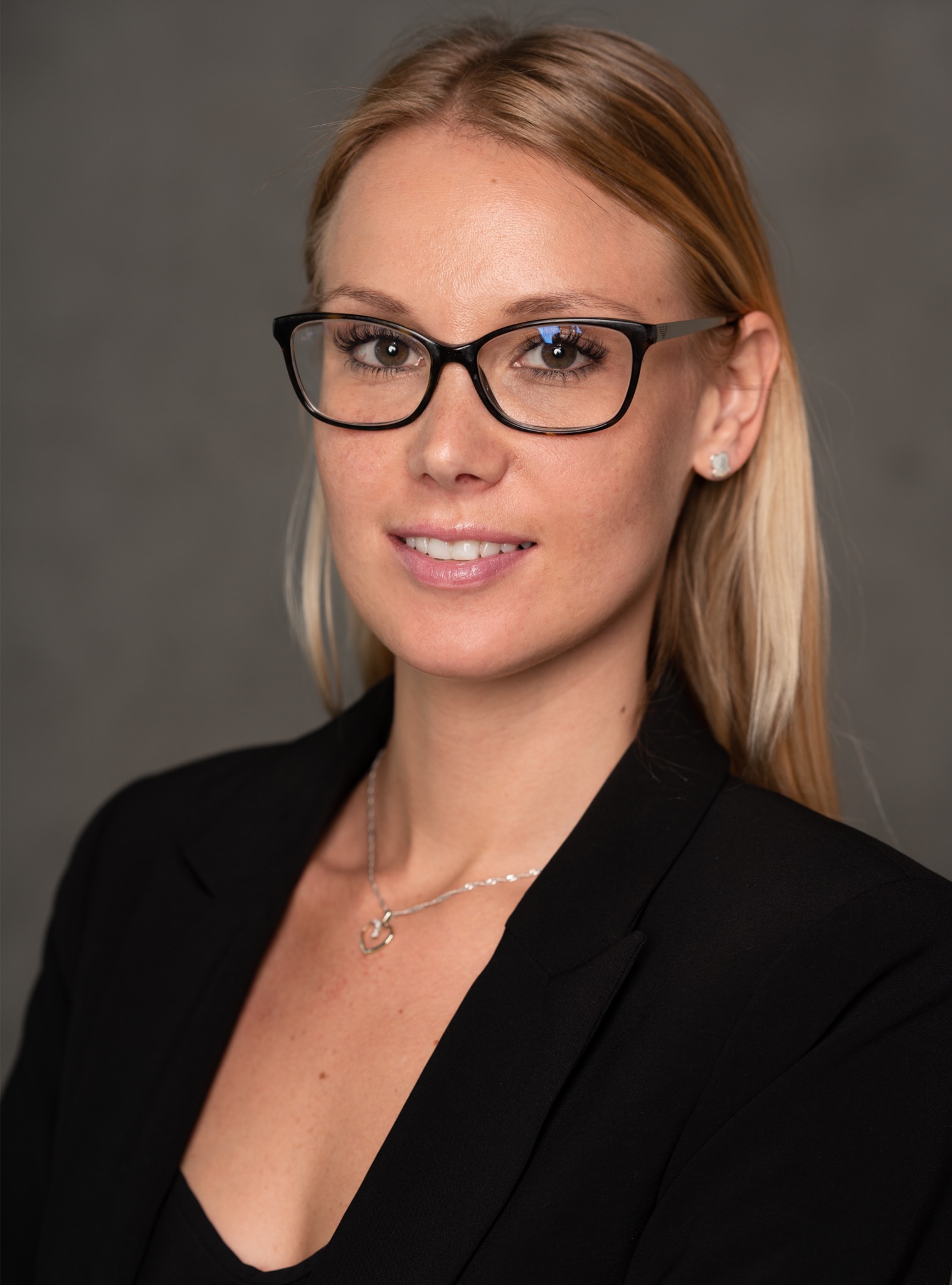 Portret uśmiechniętej blondynki w czarnej marynarce i okularach, z delikatnym naszyjnikiem na tle szarego tła.