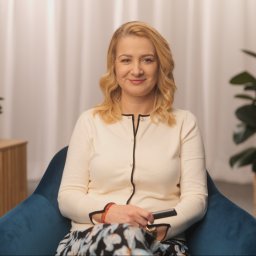 NAWA Film - Kobieta z blond włosami siedzi w niebieskim fotelu, trzymając czarny pilot w dłoni, w tle widoczny stolik z lampą i rośliny doniczkowe.