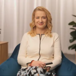 Kobieta z blond włosami siedzi w niebieskim fotelu, trzymając czarny pilot w dłoni, w tle widoczny stolik z lampą i rośliny doniczkowe.