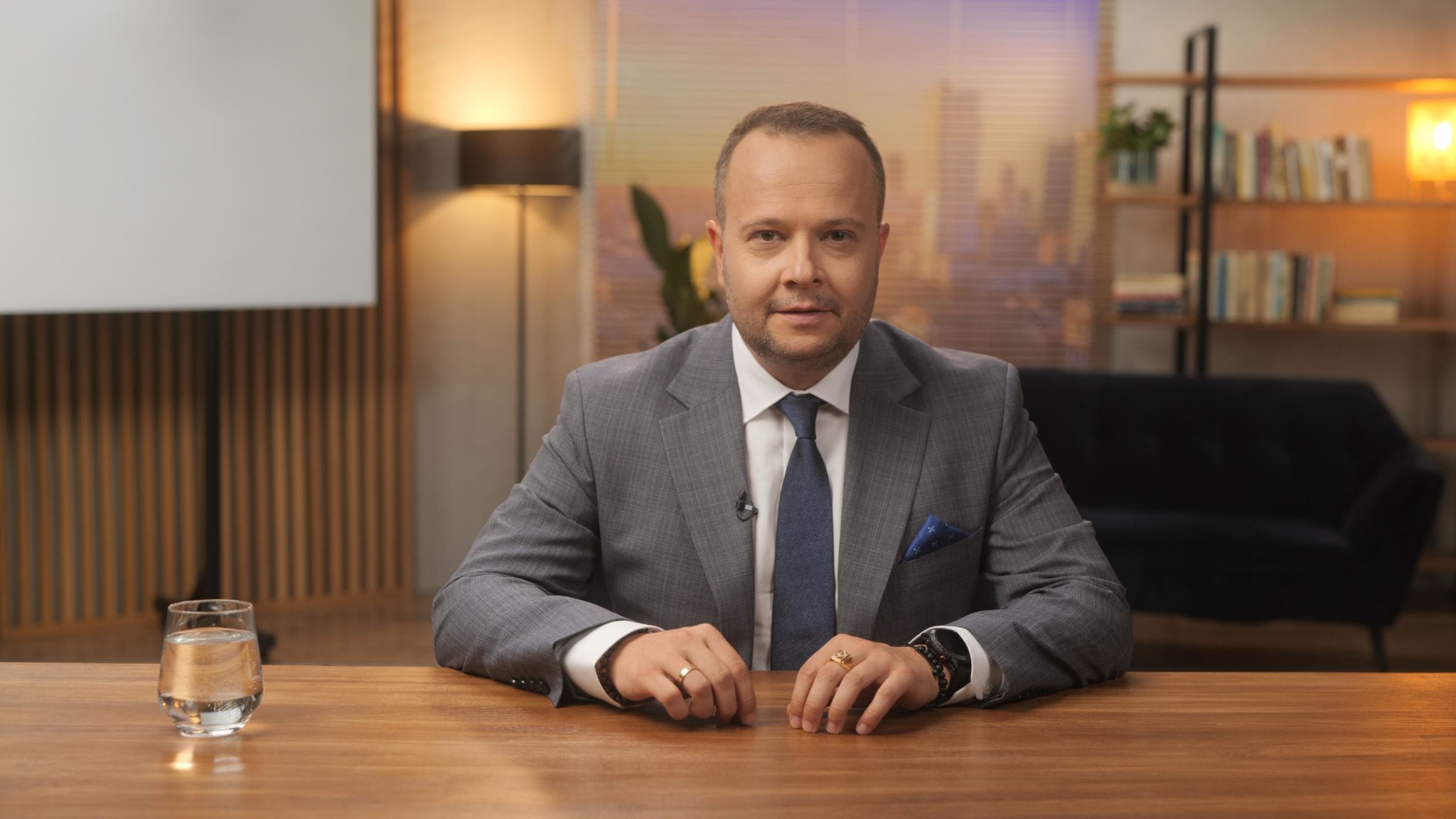 Mężczyzna w szarym garniturze siedzi za biurkiem z mikrofonem krawatowym, w tle widoczny ekran projekcyjny i regał z książkami.