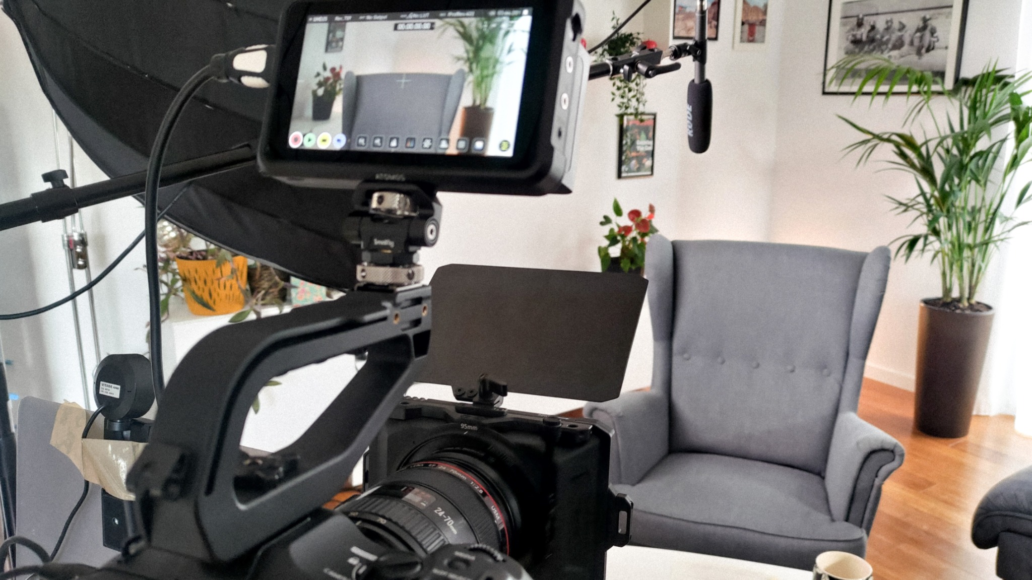 Profesjonalny sprzęt filmowy, w tym kamera z obiektywem 24-70mm, monitor podglądowy i mikrofon Rode, ustawiony w pomieszczeniu z szarym fotelem typu uszak i roślinami doniczkowymi, gotowy...