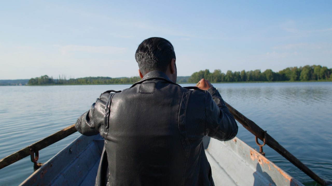 Mężczyzna w skórzanej kurtce wiosłuje łódką po spokojnym jeziorze w słoneczny dzień, widok zza pleców na oddalający się brzeg z drzewami.