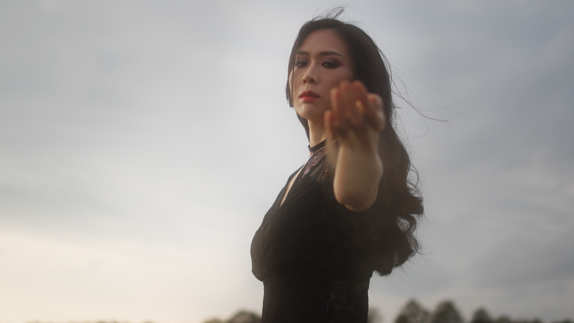 Kobieta w czarnej koronkowej sukience z wyciągniętą ręką, makijaż z czerwonymi ustami, na tle pochmurnego nieba i drzew w oddali.