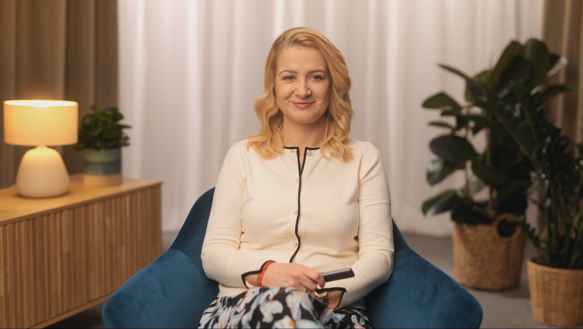 Kobieta z blond włosami siedzi w niebieskim fotelu, trzymając czarny pilot w dłoni, w tle widoczny stolik z lampą i rośliny doniczkowe.