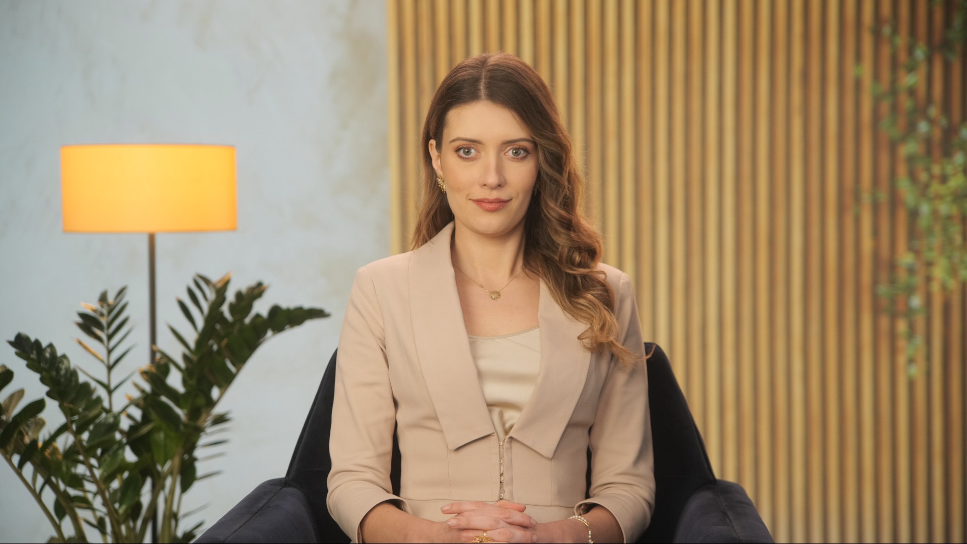 Kobieta w beżowym żakiecie siedzi na krześle na tle drewnianej ściany i rośliny doniczkowej, prawdopodobnie podczas nagrywania wideo.