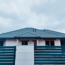 Rafdach - Dom jednorodzinny z szarą dachówką karpiówką i nowoczesnym ogrodzeniem panelowym. Widok z zewnątrz, pochmurne niebo w tle.