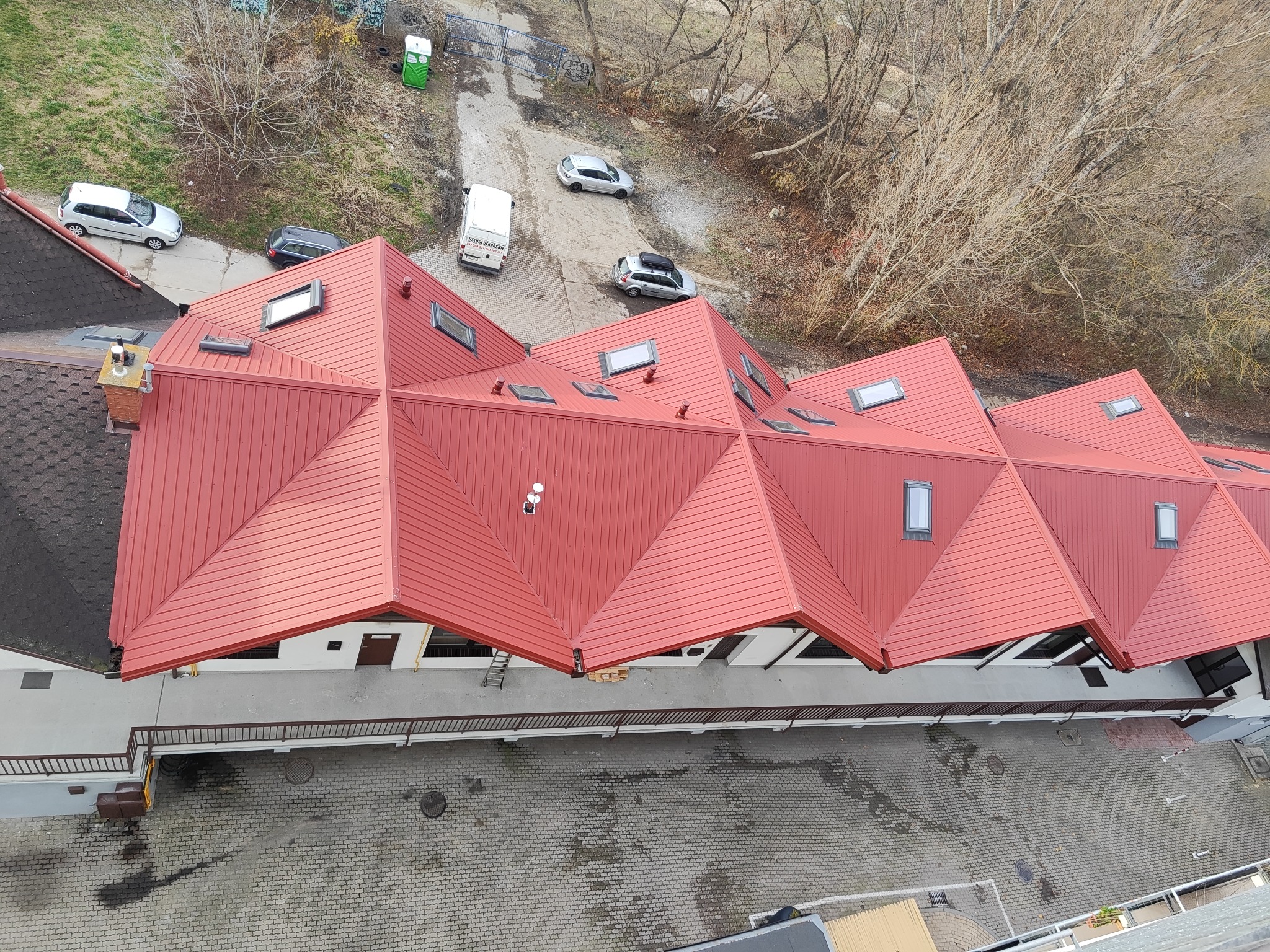 Czerwony, metalowy dach wielospadowy z oknami dachowymi, widziany z góry. Na dole budynku widoczny chodnik z kostki brukowej i zaparkowane samochody.
