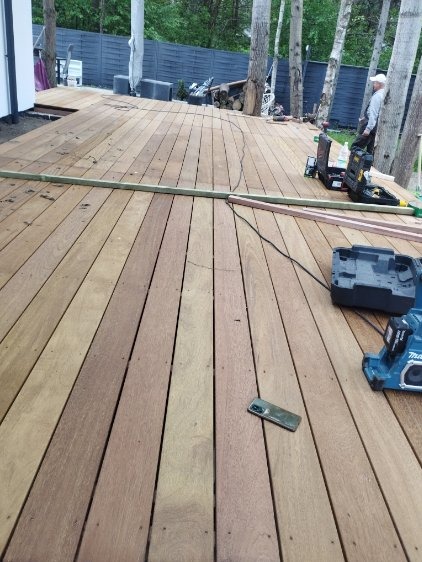 Drewniany taras w trakcie budowy, widoczne narzędzia i telefon na deskach. Częściowo ukończona konstrukcja z widocznym fragmentem ogrodzenia w tle.