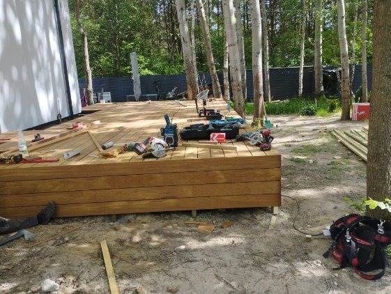 Budowa drewnianego tarasu z desek o ciepłym odcieniu brązu, otoczonego brzozowym lasem. Widoczne narzędzia i materiały budowlane w trakcie montażu.