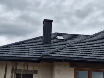 Grafitowy dach z blachodachówki modułowej, nowy komin i okno dachowe na tle pochmurnego nieba. Widoczne fragmenty elewacji w trakcie prac wykończeniowych.